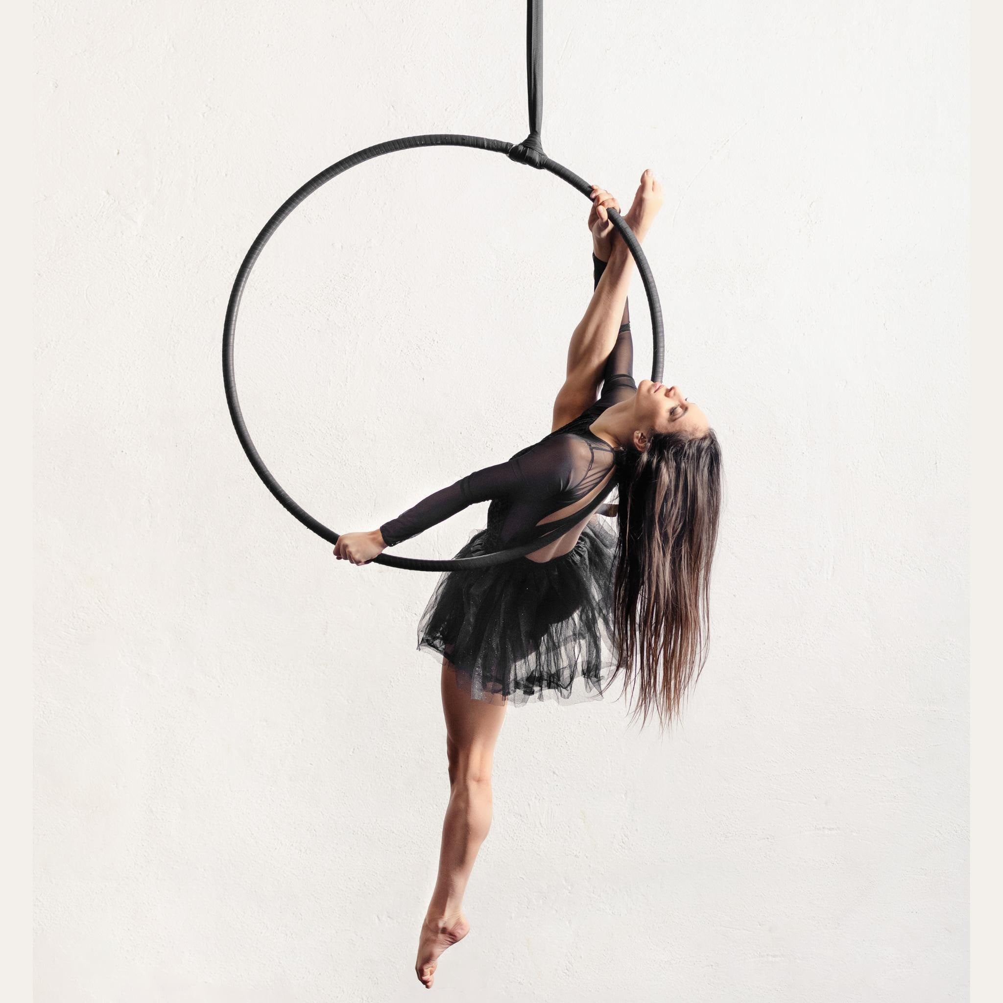 A woman in a black aerial dance leotard performs a pose on a black aerial hoop against a plain white wall.
