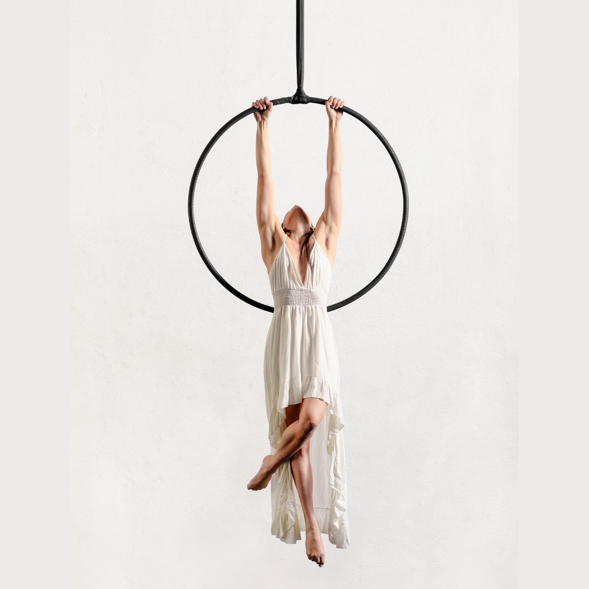 A woman in a flowing white dress hangs from a black aerial hoop against a plain light background.